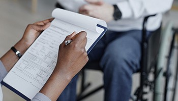hands writing on papers on clipboard sitting in front of a person in a wheelchair