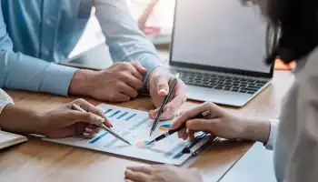 Two people reviewing financial documents and charts at a desk with a laptop nearby.