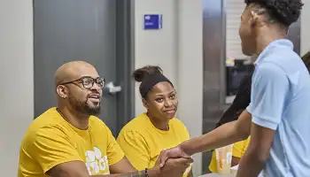 •	Two people at a welcome table shaking hands with a young adult attendee