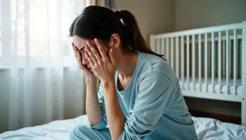 Woman sitting on bed with head in hands, in grief, with crib visible in background.