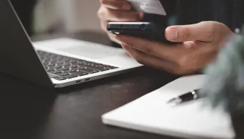 Person holding smartphone while working at laptop with notebook nearby.