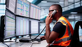 Data center worker in orange safety vest studying data on multiple computer monitors at desk.