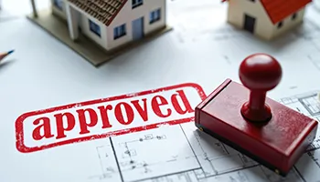 Red "Approved" stamp on architectural blueprint with house model in background.