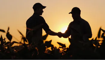 Two people shaking hands in a cornfield at sunset, silhouetted against golden sky.
