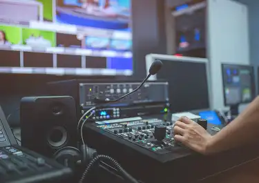 Audio engineer adjusting mixing console controls in professional studio with monitors in background.