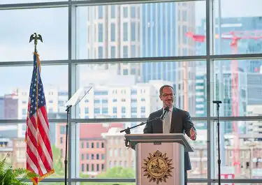Mayor Freddie O'Connell at podium with American flag, city skyline visible through windows behind him