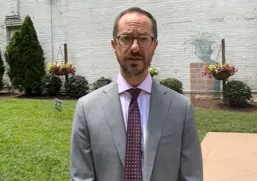 Man in gray suit and purple tie standing outdoors with brick building and green lawn behind him.