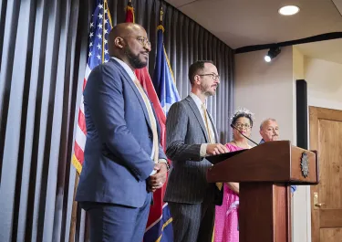 Man in grey suit and speaking at podium with American flag and colleagues nearby.