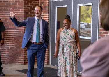 Man in blue suit waving at woman in floral dress outside brick building entrance.