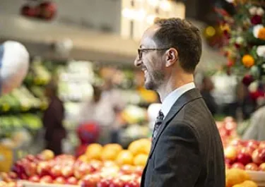 Man in glasses and dark suit standing in a vibrant produce market with colorful fruits and vegetables in background.