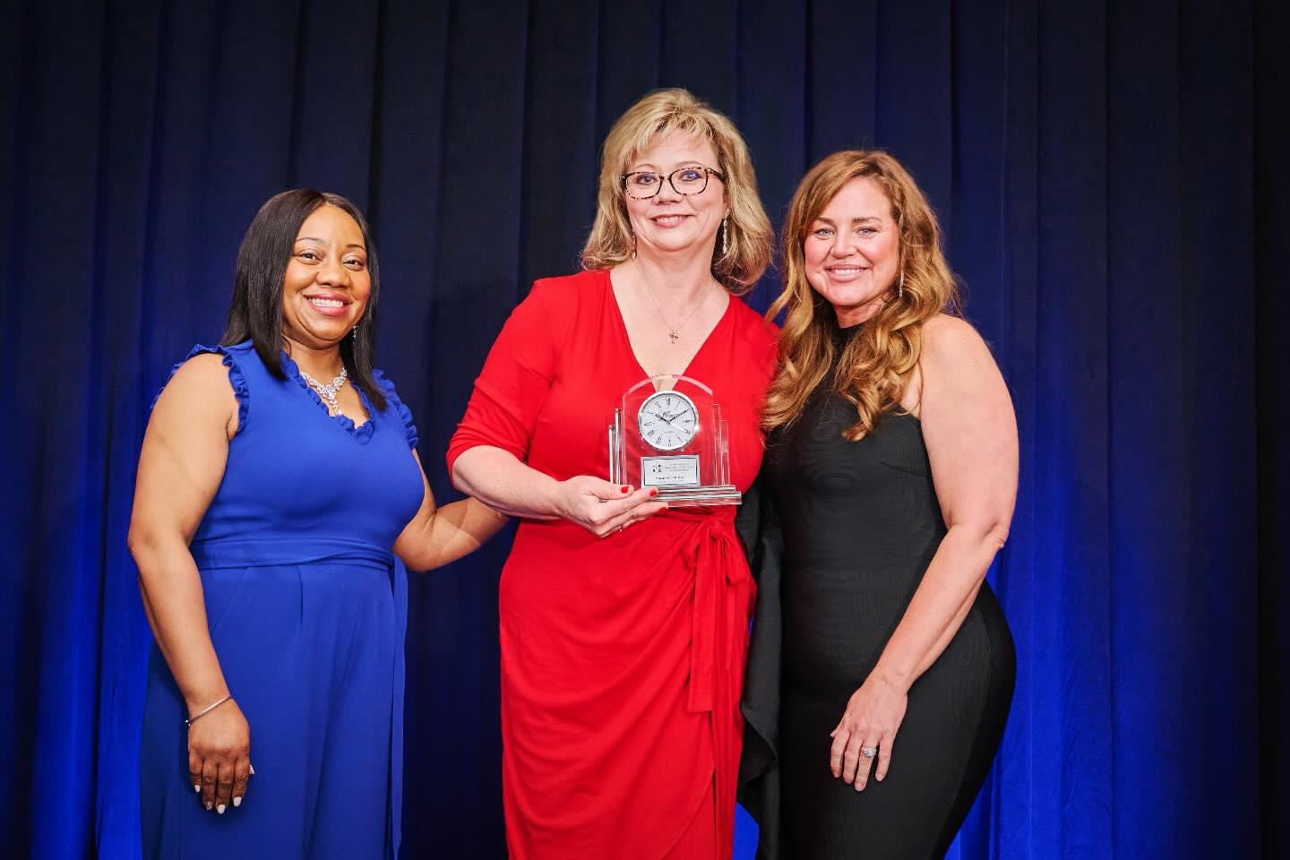 Three women standing together against a blue curtain backdrop, with the center woman in red holding an award.