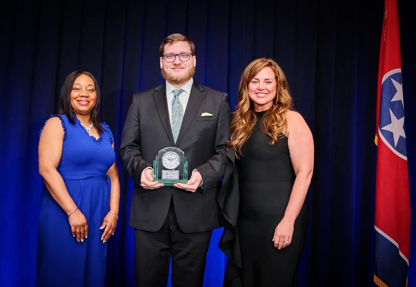 Three people in formal attire standing in front of flags and blue backdrop, man in center holding an award.