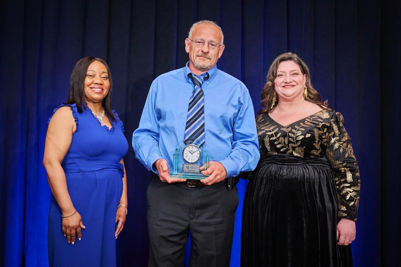 Three people standing together on stage in formal attire against a blue curtain backdrop.