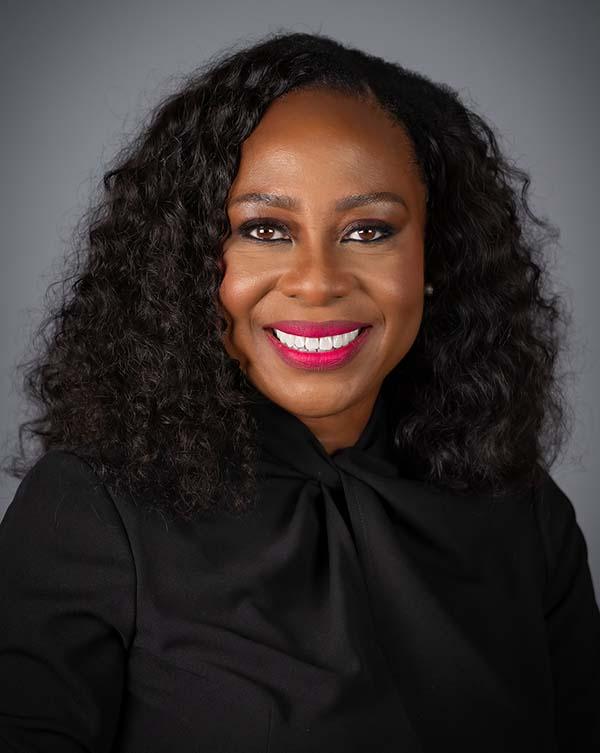 Woman with long dark curly hair wearing black shirt, smiling at camera against gray background.