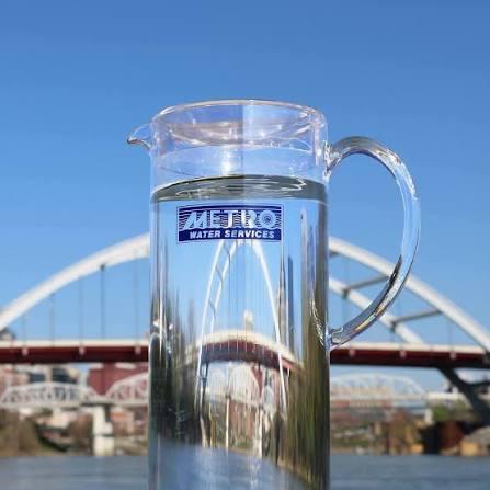 Clear pitcher of water against blue sky with bridge reflected in it.