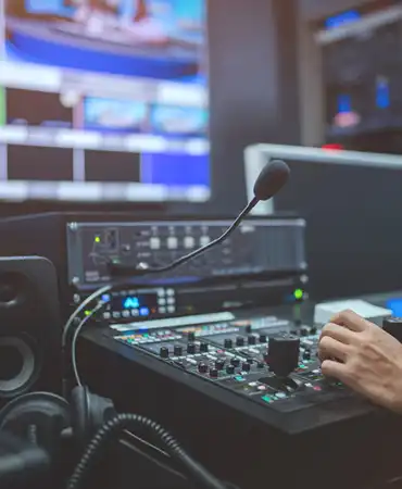 Audio engineer adjusting mixing console controls in professional studio with monitors in background.