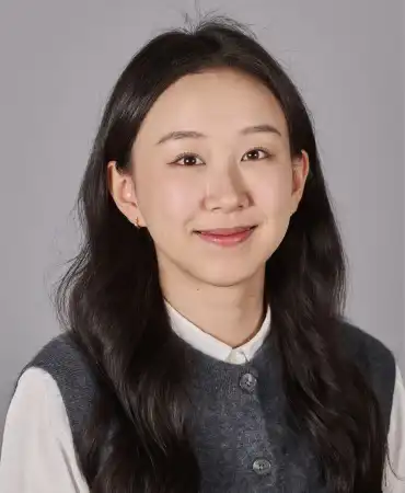 Young woman with long dark hair wearing a dark vest over white shirt, smiling at camera against gray background.