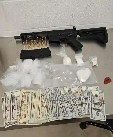 Black rifle with ammunition, magazines, and cash laid out on white surface during police seizure.