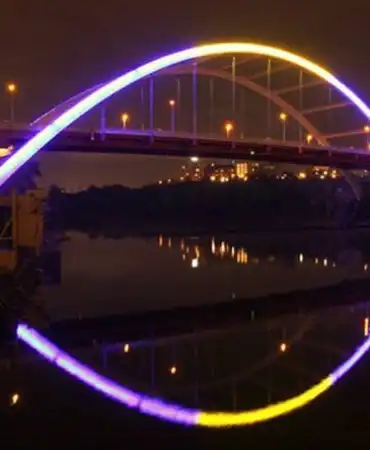 Illuminated circular arch bridge reflecting in dark water at night with purple and yellow lights.