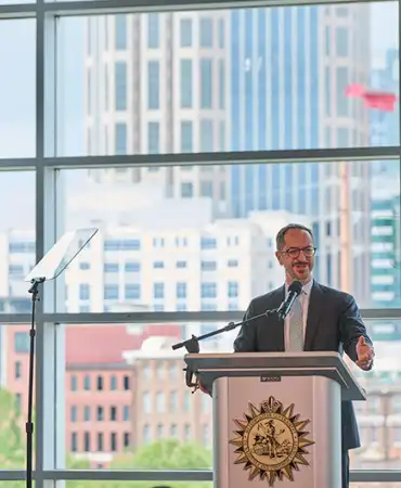 Mayor Freddie O'Connell at podium with American flag, city skyline visible through windows behind him