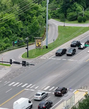 Aerial view of a busy urban intersection with multiple vehicles, traffic lights, and green landscaping on both sides of the road.