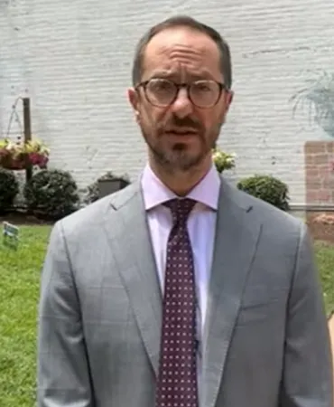 Man in gray suit and purple tie standing outdoors with brick building and green lawn behind him.