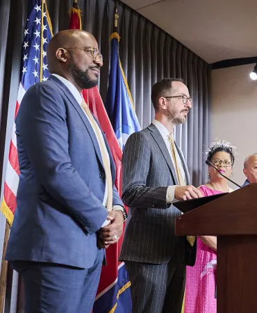 Man in grey suit and speaking at podium with American flag and colleagues nearby.