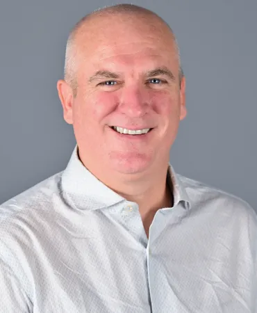 man with friendly smile wearing light gray button-up shirt against gray background.