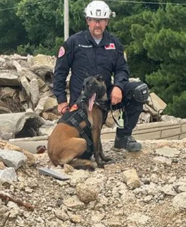 Person in protective gear sitting on rocky ground with brown dog beside them.