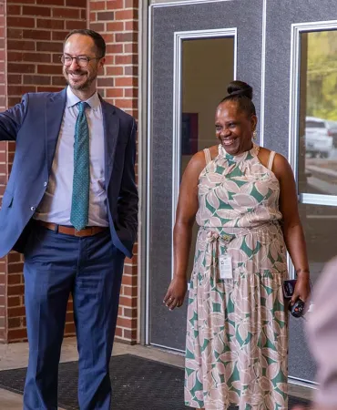 Man in blue suit waving at woman in floral dress outside brick building entrance.