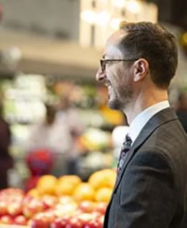 Man in glasses and dark suit standing in a vibrant produce market with colorful fruits and vegetables in background.