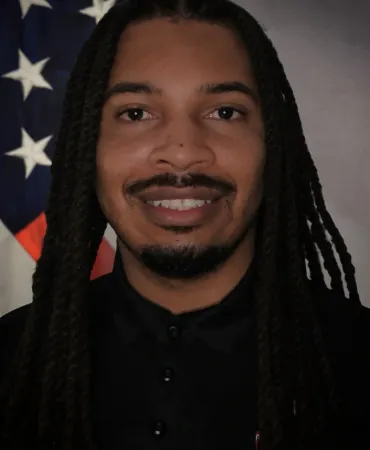 Man with long dreadlocks smiling at camera with American flag in background.