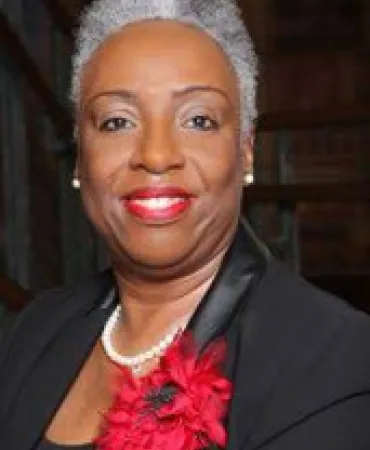 Professional headshot of a woman with white hair, wearing a black blazer and bright red flower brooch.