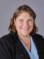 Professional headshot of a smiling woman with brown hair wearing a dark blazer and blue shirt.