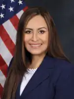 Professional portrait of a woman in dark blazer with American flag background.