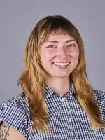 Woman with long blonde hair wearing a blue checkered shirt, smiling at camera against gray background.