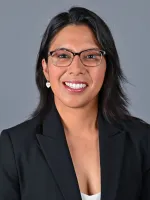 Professional headshot of a smiling woman wearing glasses and a dark blazer against a gray background.