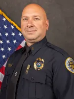 Police officer in dark uniform standing in front of American flag, official portrait photograph.