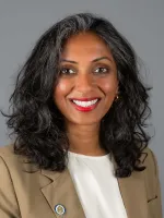 Woman with long black wavy hair and red lipstick wearing a tan blazer over white shirt, professional headshot.