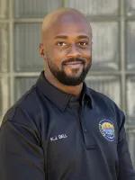 Smiling man in dark blue polo shirt with emblem against industrial window backdrop.