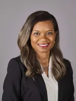 Professional headshot of a woman with long dark hair wearing a black blazer and white shirt, smiling at camera against gray background.