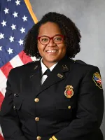 Woman in fire department dress uniform wearing glasses, with American flag in background.