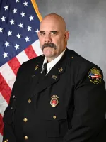 Bald man with mustache in dark uniform with medals standing in front of American flag.