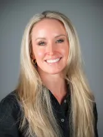 Professional headshot of a smiling woman with long blonde hair wearing a dark blazer against a gray background.